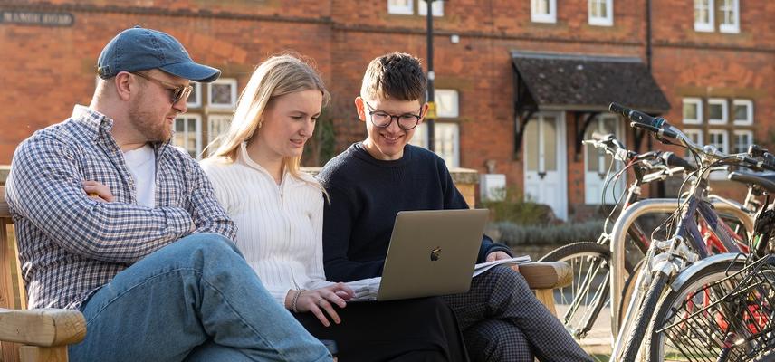 Three undergraduates sitting on a bench looking at a laptop computer